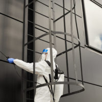 Wide angle view of one worker wearing protective suit spraying chemicals over black building facade during disinfection or cleaning, copy space