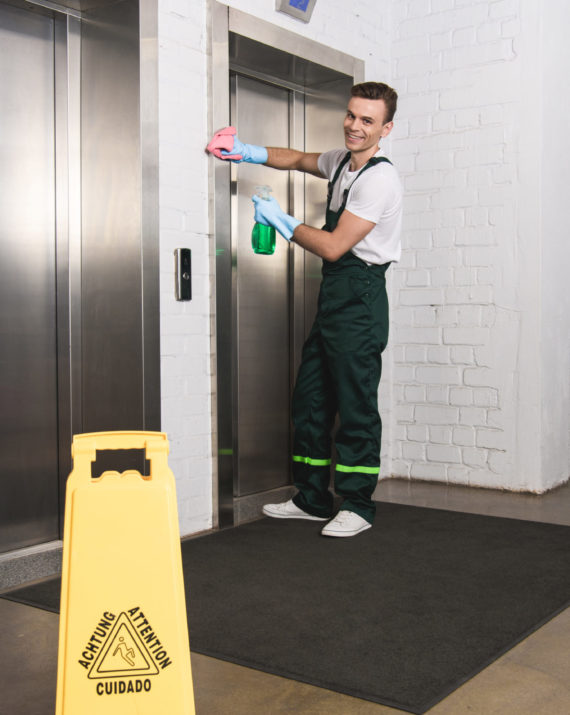 handsome young janitor cleaning elevator and smiling at camera